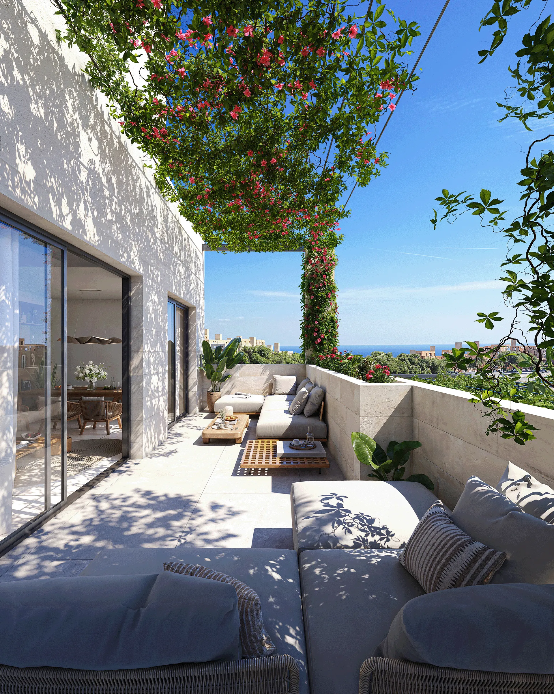 Sunlit terrace with cushioned seating, wooden tables, plants, flowering vine canopy, and ocean in the distance.